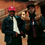 Two men in jackets and caps stand in a dusty outdoor yard beside a brick building with a stairway in the background; caption reads 'W'abre a, bra fie (Bo Bo Bo Bo)'