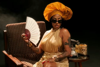 Elegant woman in a gold dress and oversized orange headwrap sits in a wicker chair, holding a white fan and wearing dark sunglasses.